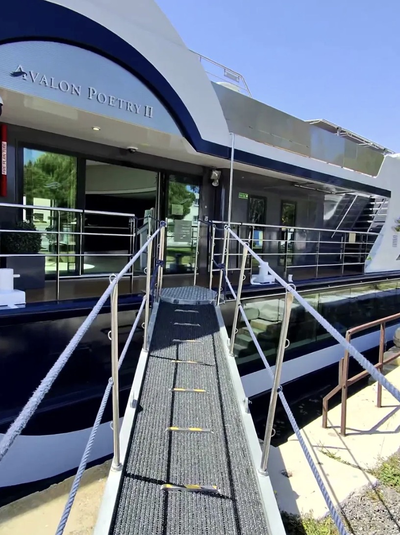 A gangway with textured non-slip treads and handrails leading up to the Avalon Poetry II, docked in port under clear blue sky.